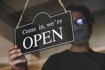 man wearing mask holds an &quot;open&quot; business sign