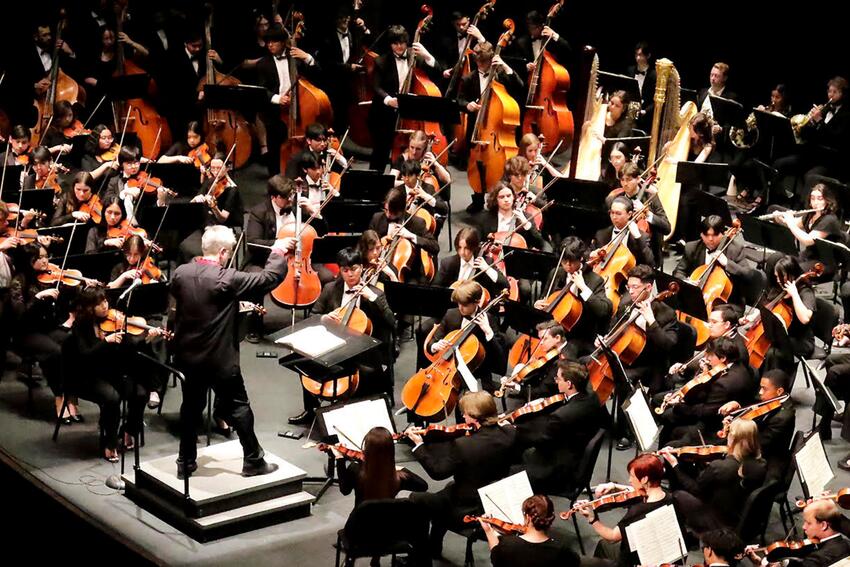 Taras Krysa, on a podium with his hands and baton raised, conducts the full UNLV Symphony Orchestra (performers and instruements are displayed in the image).