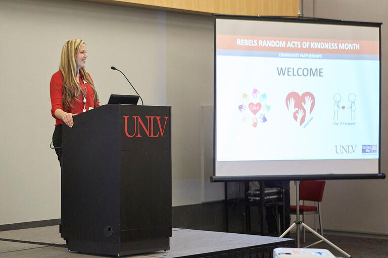 A woman stands at a podium, speaking to an unseen audience. An image is displayed on a screen to her right.