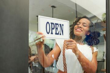 smiling black woman hangs &quot;open&quot; sign on the window of her business