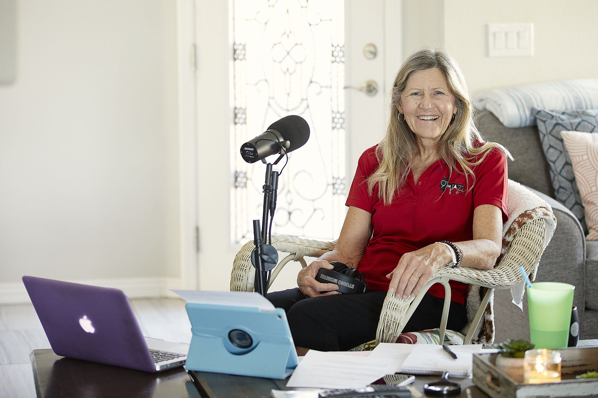 A woman sitting in a living room with a microphone and various tablet devices on the table.