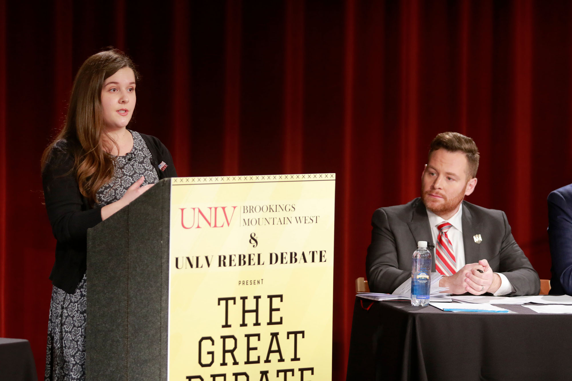 Woman at podium that says &quot;The Great Debate&quot; talking while man listens from table