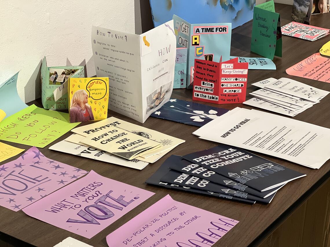 A table is covered with paper signs and small publications. Some of them are obviously handmade, with text written in felt-tip pen or spelled out in glued-on cut-out letters. They all have headings or titles related to voting or societal change, like, “How to Change the World,” “Many Voices. One Shared Purpose. The Right to Vote!” and “Democratize your Commute.”