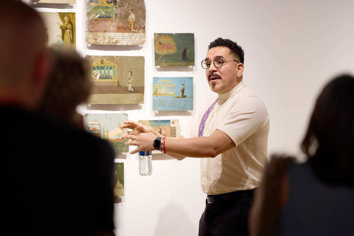 We are looking through a crowd of heads at a well-dressed man with short curly black hair who holds out his hands expressively and opens his mouth to make a (presumably important) point as he stands in front of a collection of small rectangular paintings hanging on a white wall. The paintings are all different, but almost every one includes a human figure kneeling next to a bearded man in a canopied bed.