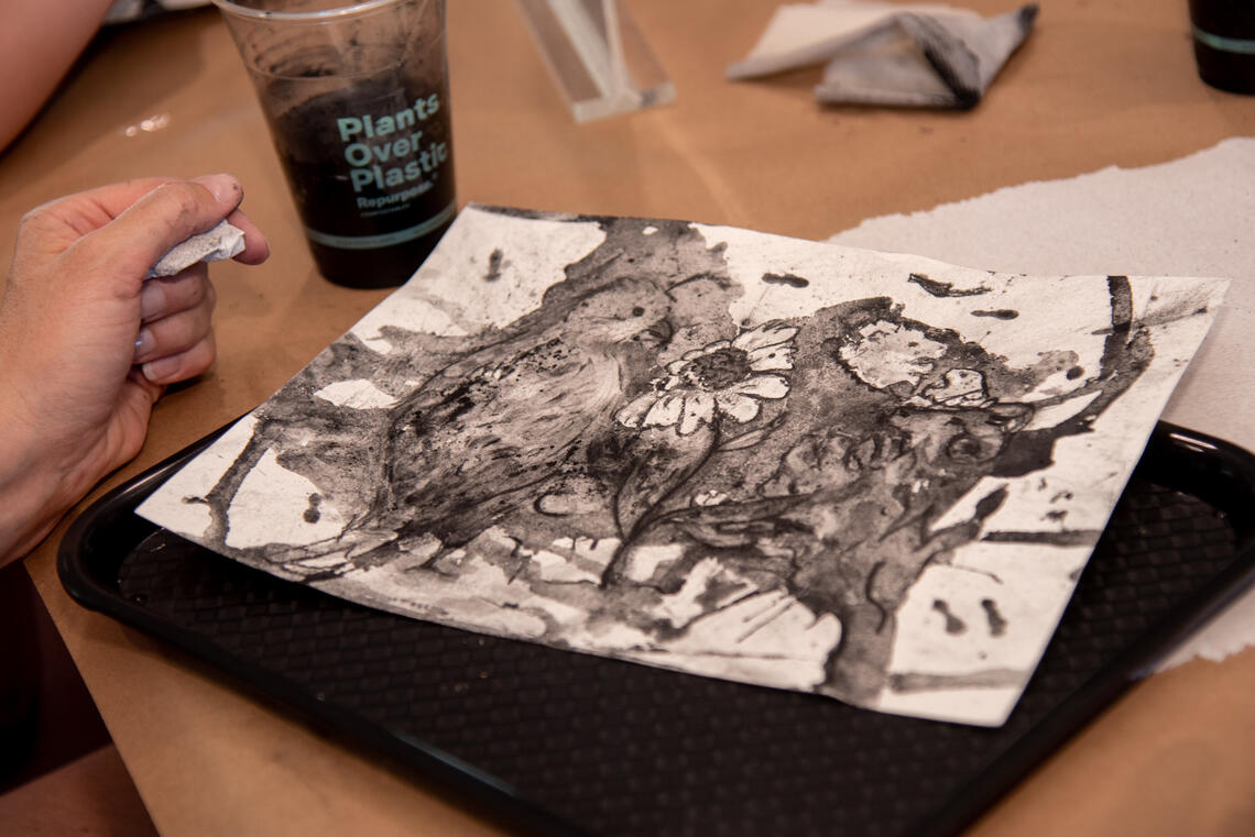 A hand holding a crumpled piece of towel hovers over a black and white drawing of a cute quail standing by a flower. The drawing is intentionally stained with puddles of black liquid but the petals of the flower are white. There's a cup of black fluid on the table next to the drawing.