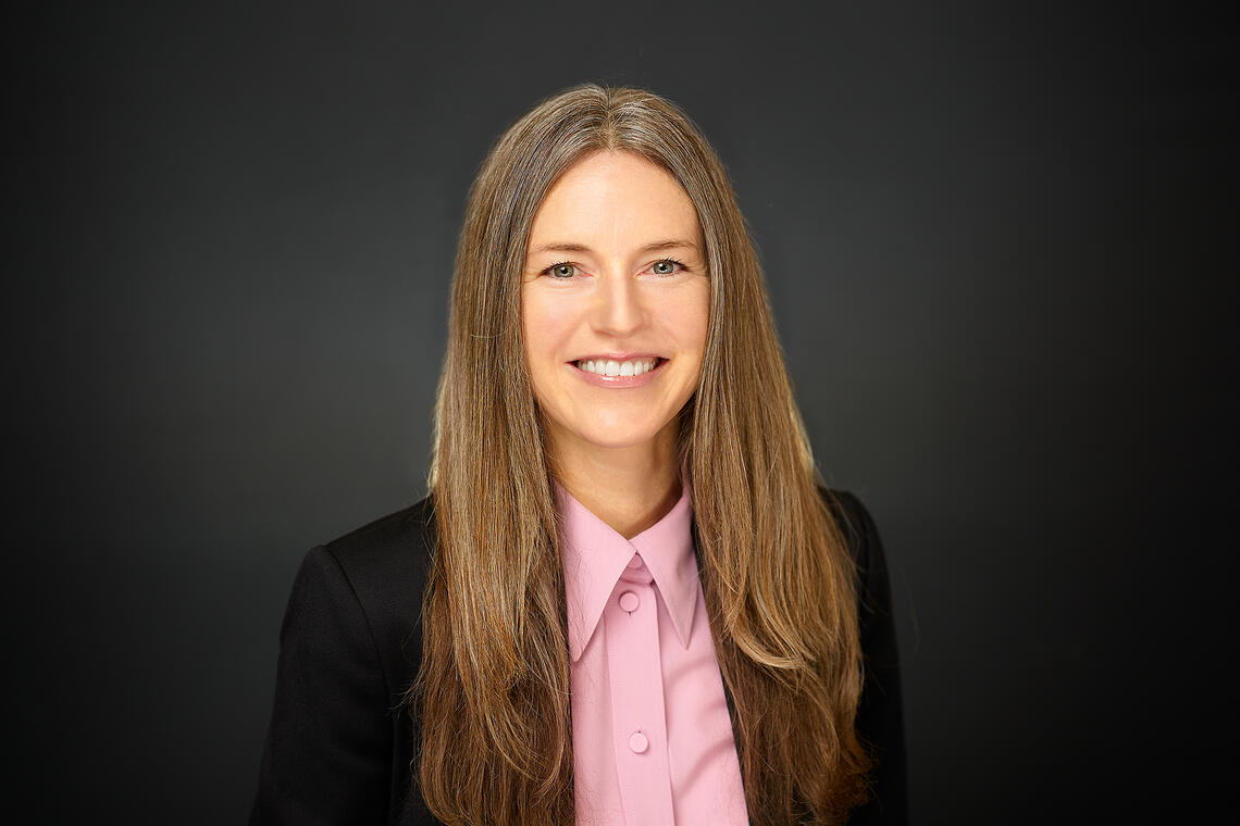 A professional headshot of a woman with long brown hair smiling at the viewer. She is dressed in a neat pink shirt and dark jacket with her hair parted cleanly down the middle.