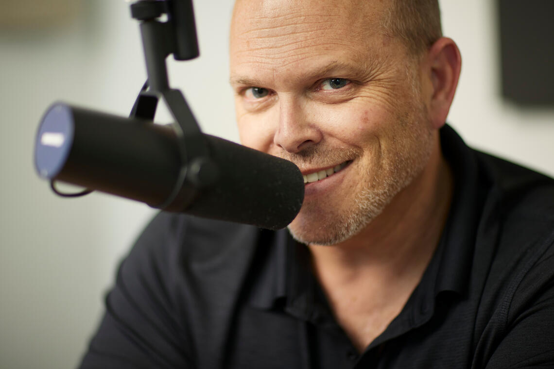 Photo of Prof. Adam Paul smiling into the camera while talking into a podcast mic