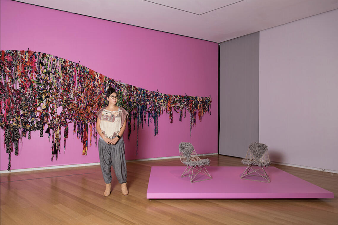 A woman in loose comfortable clothing stands with her hands together in front of a deep pink-magenta wall. She is looking off to the side. The curve of a huge artwork made from hanging, knotted, tasseled strips of patterned cloth swoops across the wall behind her head. Two gray chairs with woven fiber backs and seats stand on a pink pedestal next to her.