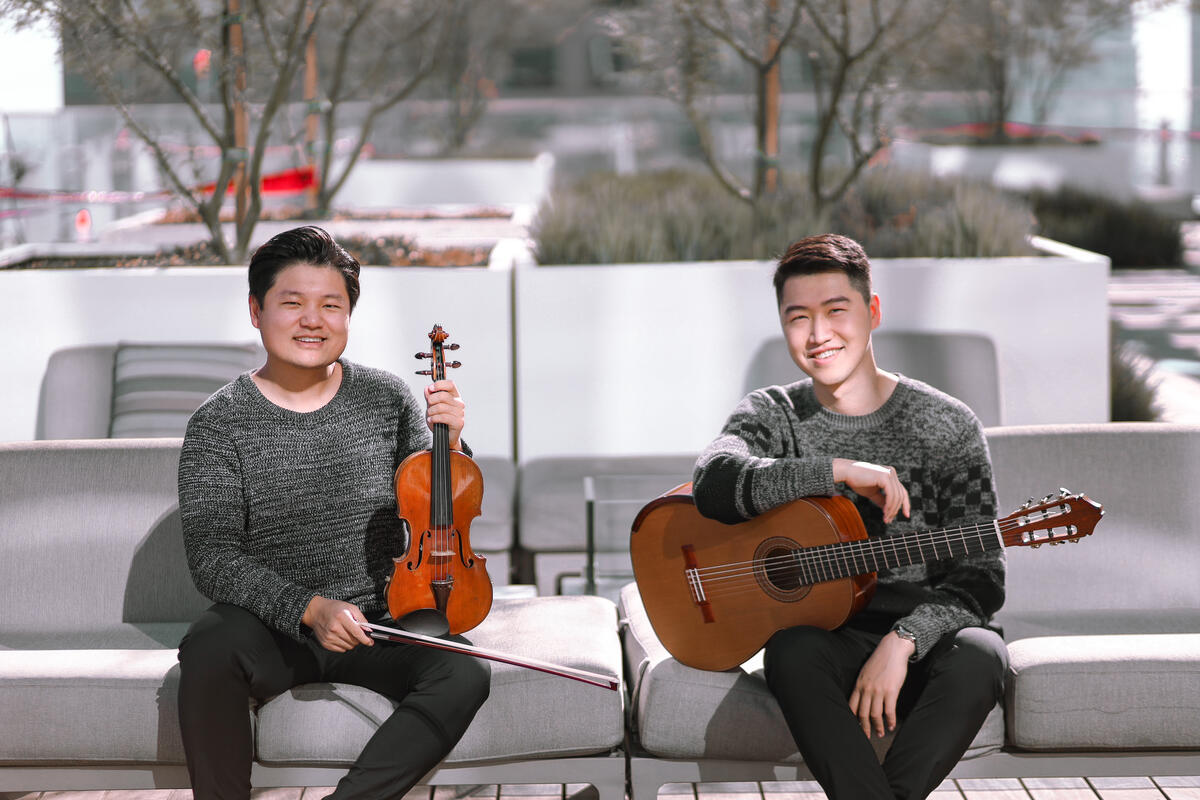 ArcoStrum, Strauss Shi (on left, holding a violin) and TY Zhang (on right, holding a guitar), looks towards the camera. They are outside on a couch in this promotional photo.