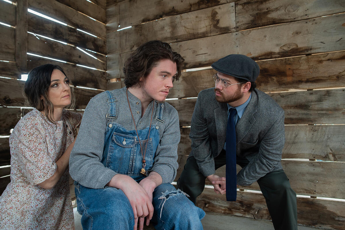 Three people sitting in a barn, two are staring at a man in the middle