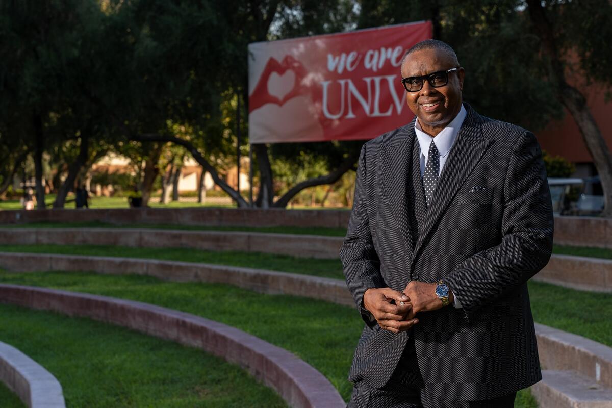 Wearing a dark suit and sunglasses, UNLV Graduate College Alumnus of the Year Lonnie Wright poses on campus with a We Are UNLV sign in the background