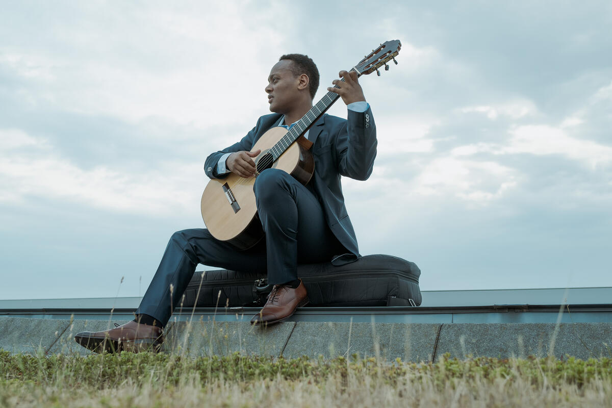 Raphael sits on a building near a field, playing his guitar.