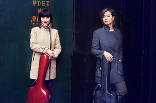 The Latin GRAMMY®-nominated Beijing Guitar Duo–Meng Su &amp; Yameng Wang–are in winter coats and looking towards the camera. Their guitar cases are in front of them.