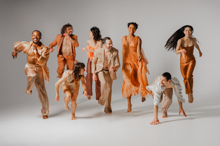 Dancers from BODYTRAFFIC, in beige and orange colored costumes, display their &quot;vivid theatricality.&quot; They're dispersed in various positions across the stage, and all are smiling. Some dancers are looking at one another and some at the camera.