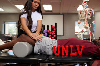 A physical therapist working with a patient. Red UNLV letters are placed on the patient's medical bed.
