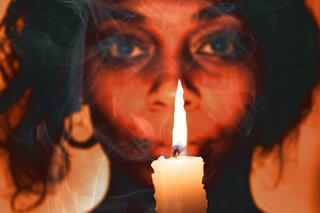 A woman (perhaps a bruja) holds a candle and looks towards the camera in this promotional image.