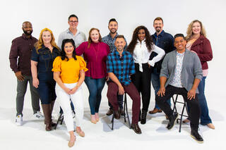 The vocal group, Voctave, with some members seated and some standing, all in casual clothes, looks toward the camera in this promotional photo.