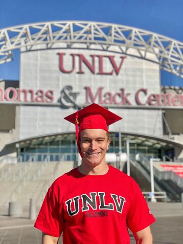 man in red graduation cap and UNLV shirt poses in front of sign that reads UNLV Thomas & Mack Center