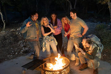 A group of actors stand around a campfire.