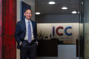 Wearing a dark blue suit and blue tie, 2024 UNLV Outstanding Alumnus of the Year Randy Garcia poses with his hands in his pants pockets in front of logo for his company, the Investment Counsel Company