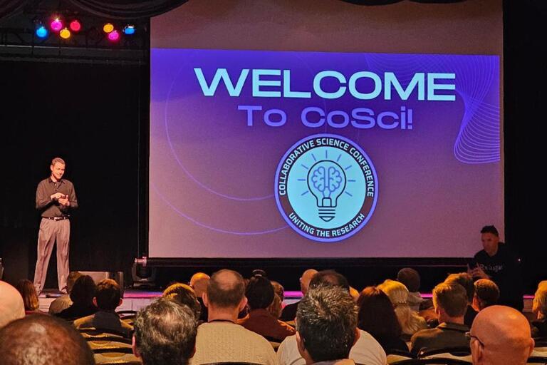 A speaker is on stage at a conference. In the photo, a big screen displays &quot;Welcome to CoSci.&quot; Conference attendees are in the audience listening to the speaker.