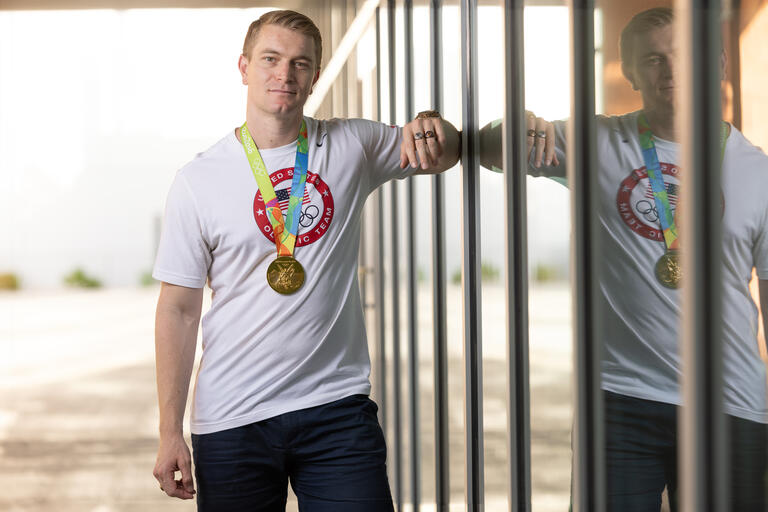 man wearing Olympics shirt and medal leans against glass wall