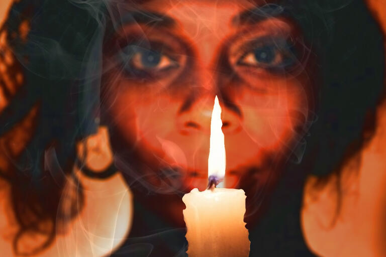 A woman (perhaps a bruja) holds a candle and looks towards the camera in this promotional image.