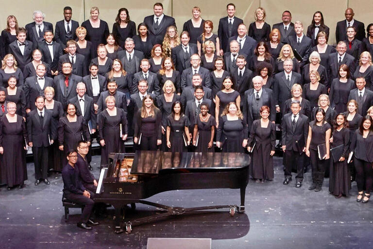 One choir in the UNLV Choral Ensembles is on stage at the Artemus W. Ham Concert Hall. A piano and pianist are in front of the ensemble. They are looking up towards the camera.