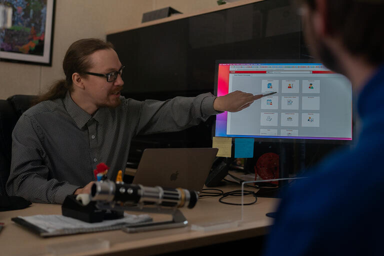 Photo of Academic Advisor speaking to student sitting in foreground. Advisor is pointing at a website on his desktop computer screen.