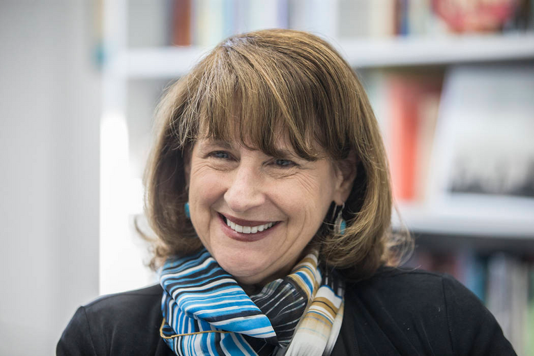 Nancy Uscher wearing a neck scarf and sitting in front of a bookcase smiling into the camera