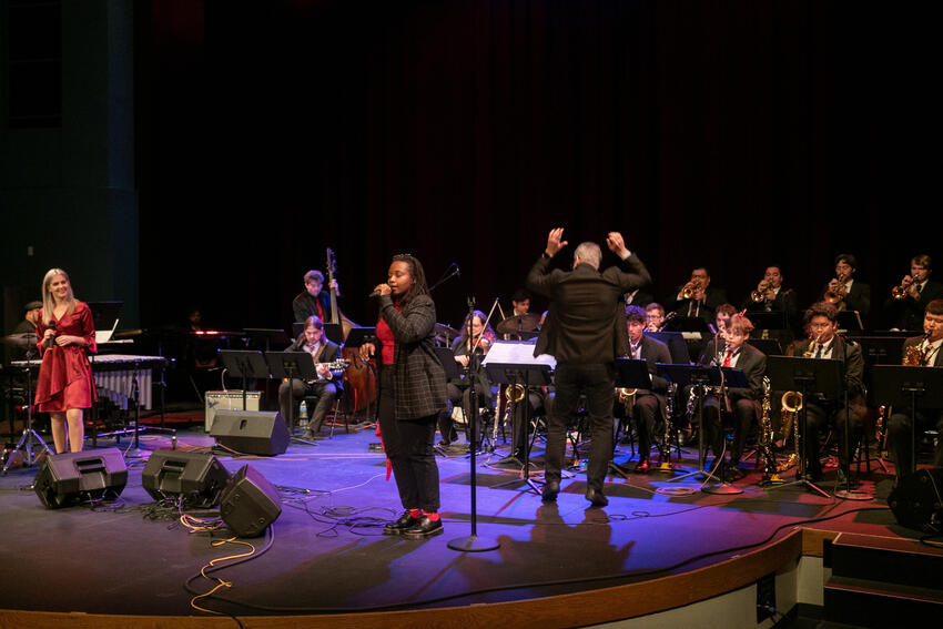 A vocalist sings as another looks on while a big band plays behind them.
