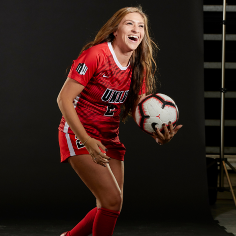 lady in a unlv soccer uniform holding a red and white soccer ball while smiling