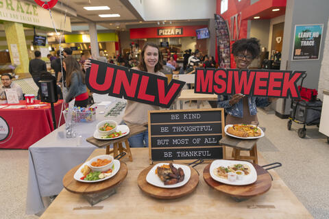 two students in a dining area holding signs that say &quot;UNLV&quot; and &quot;MSI WEEK&quot;